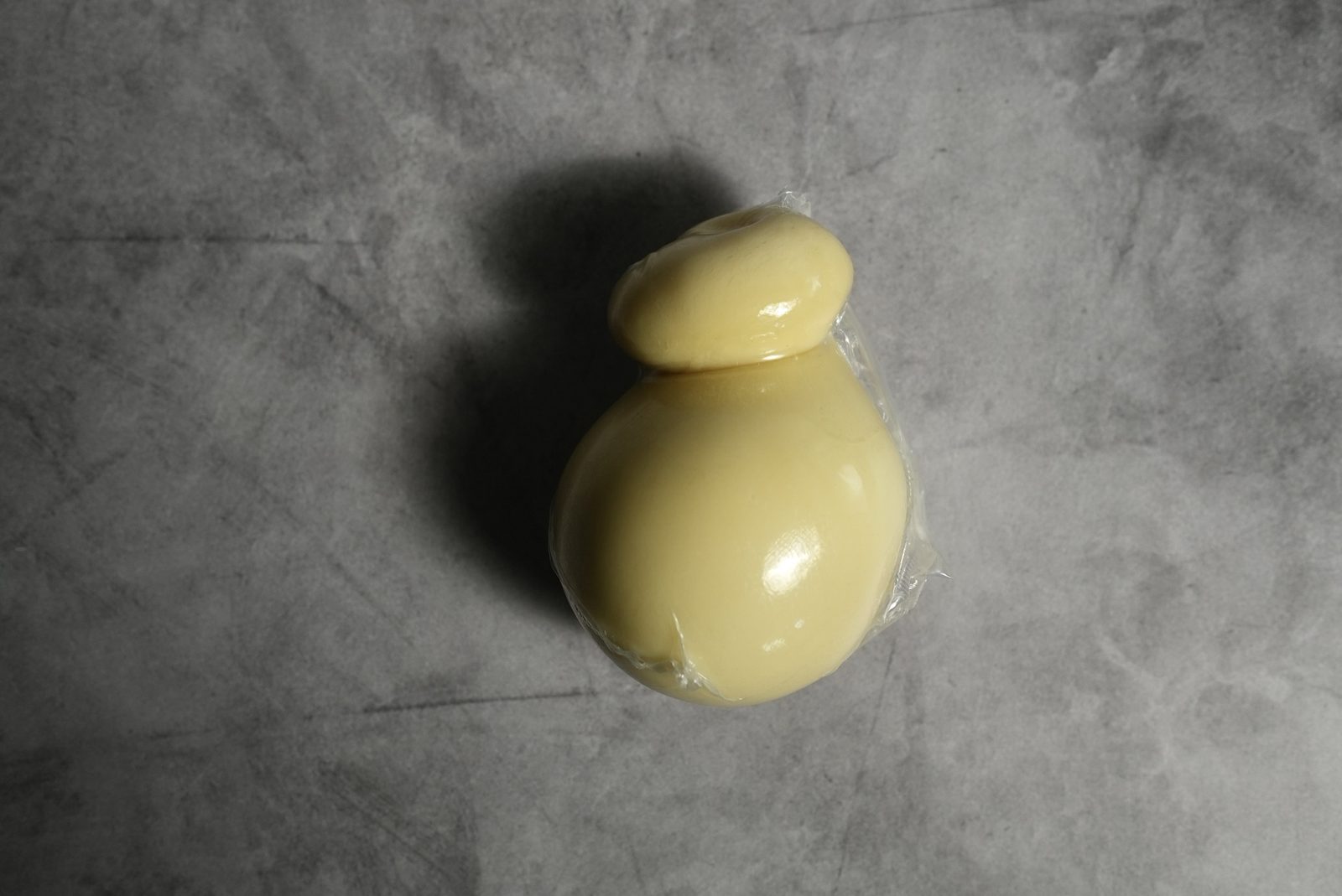 A pear-shaped scamorza with its tied knot, wrapped, on a gray surface.
